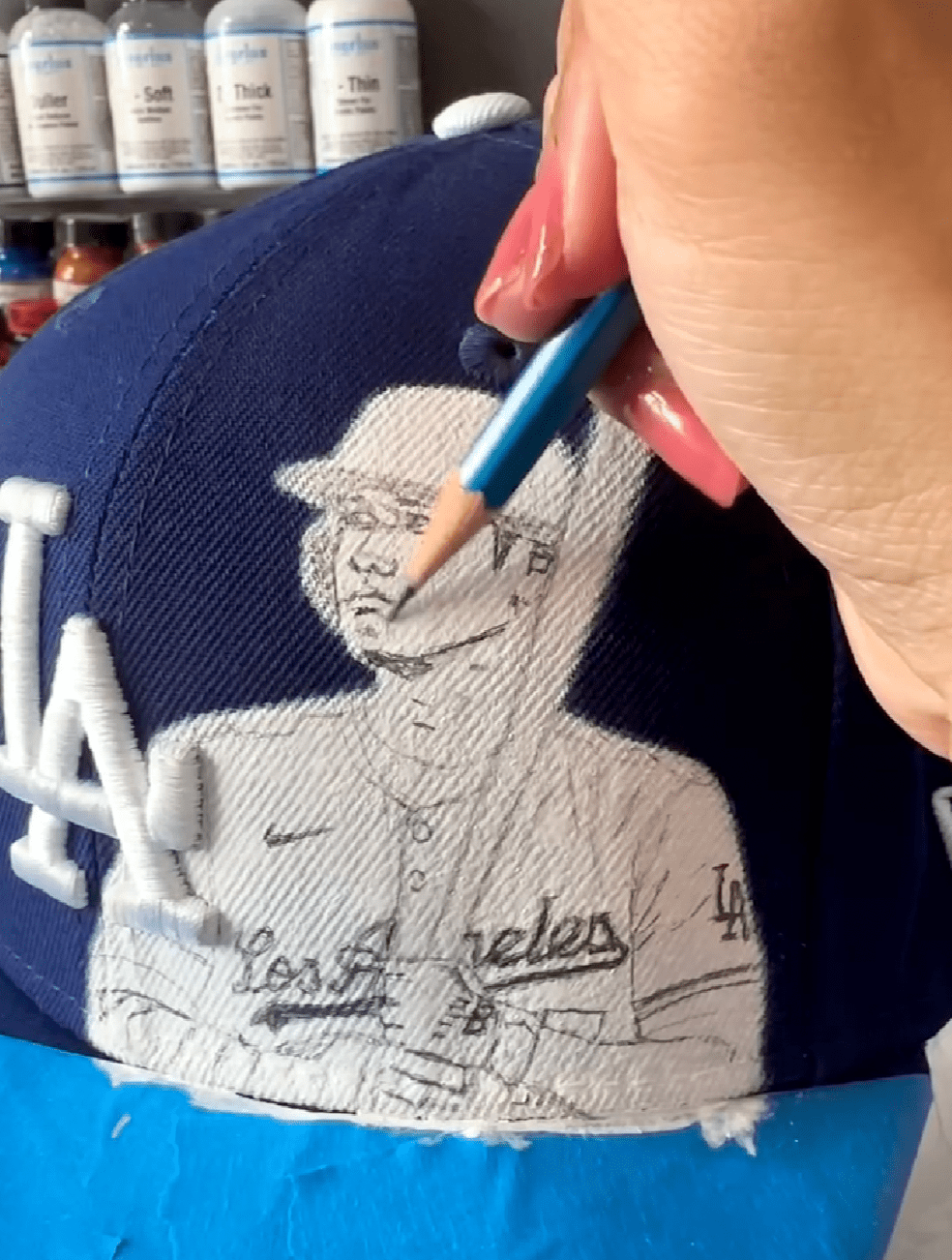 Image displays a pencil sketch lightly drawn onto the curved fabric surface of a hat, outlining the planned design guides for painting.
