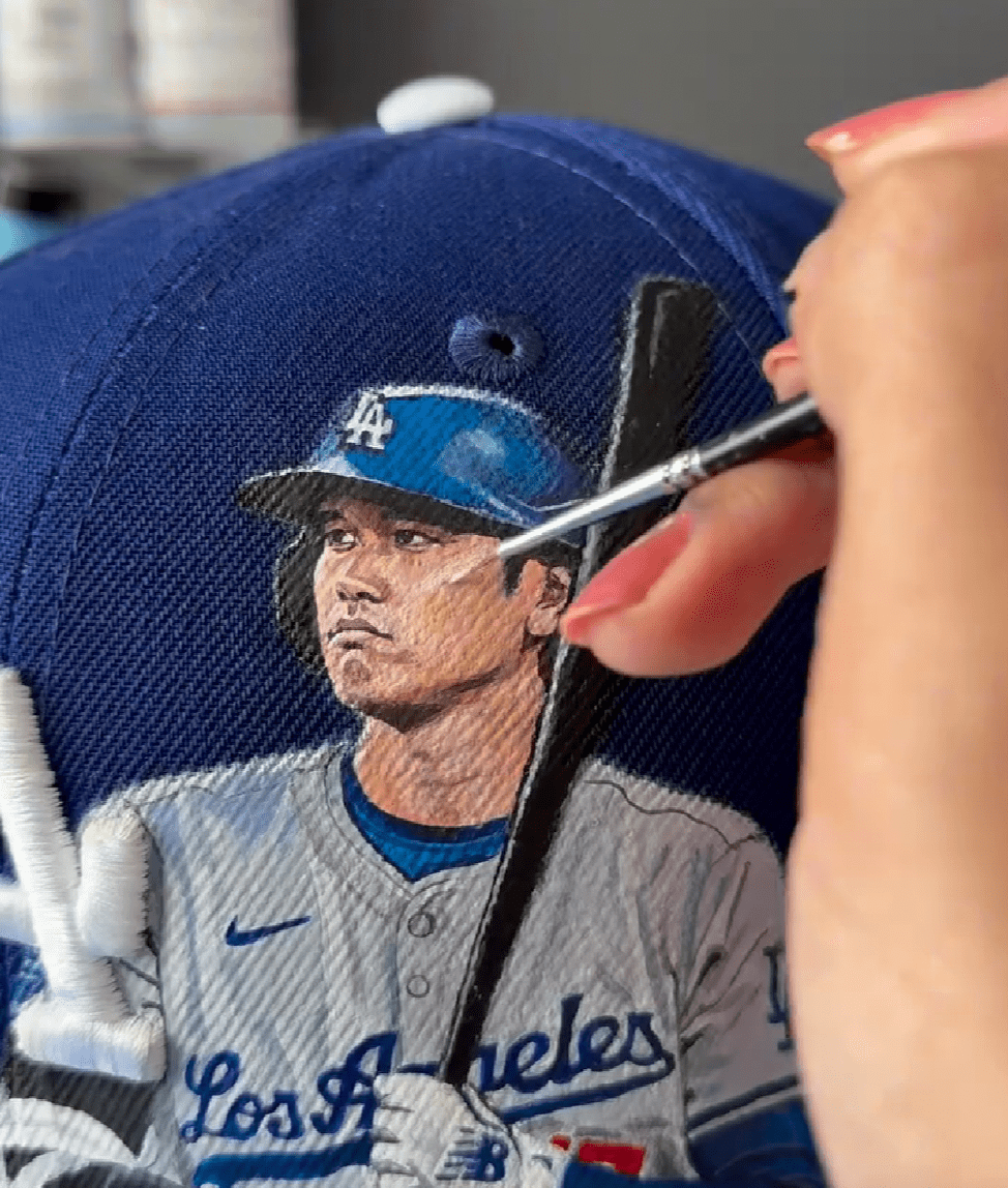 Photo shows artist painting very detailed image of baseball player, Shohei Ohtani onto a baseball cap.