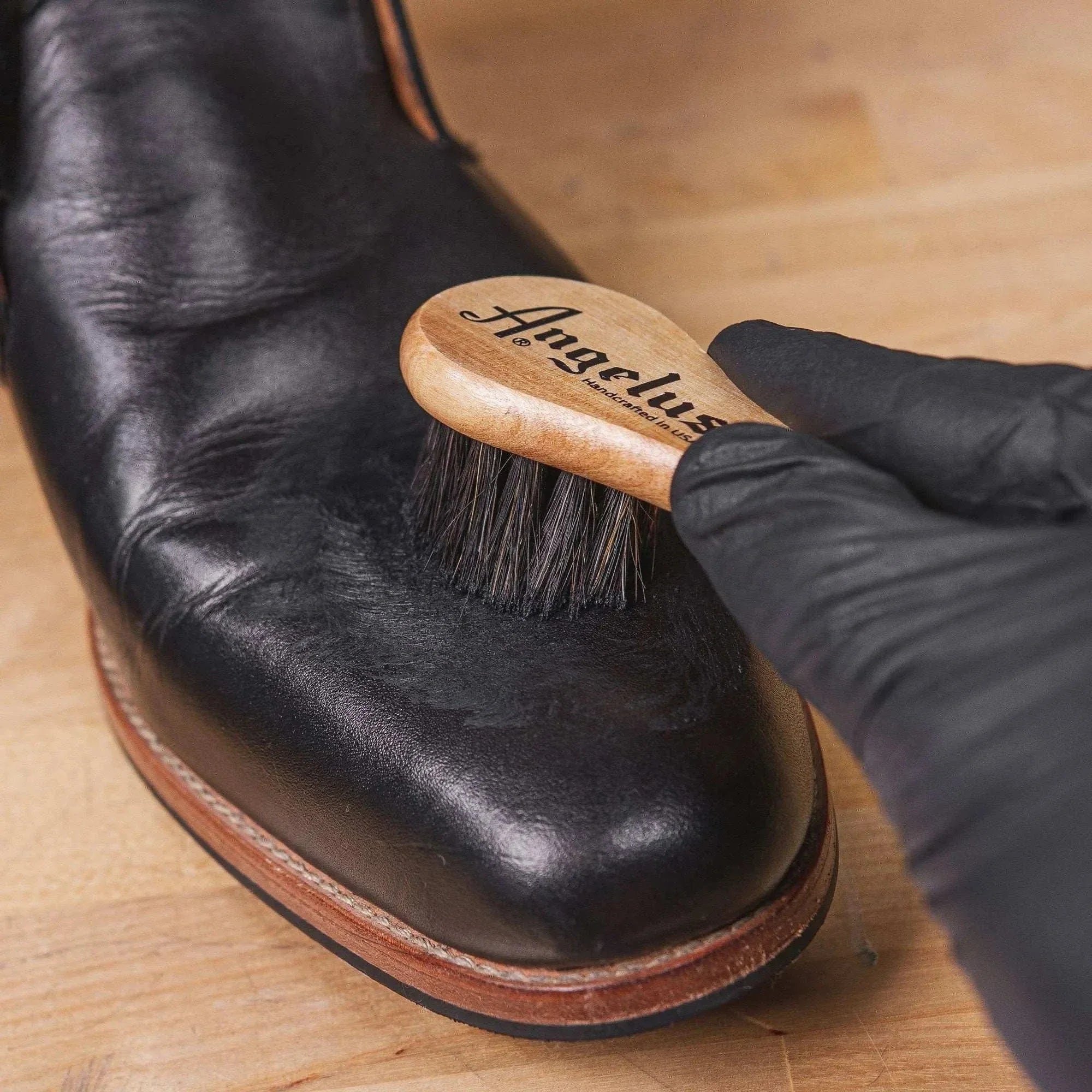Gloved hand brushing a black leather boot with an Angelus brush to apply leather boot conditioner.