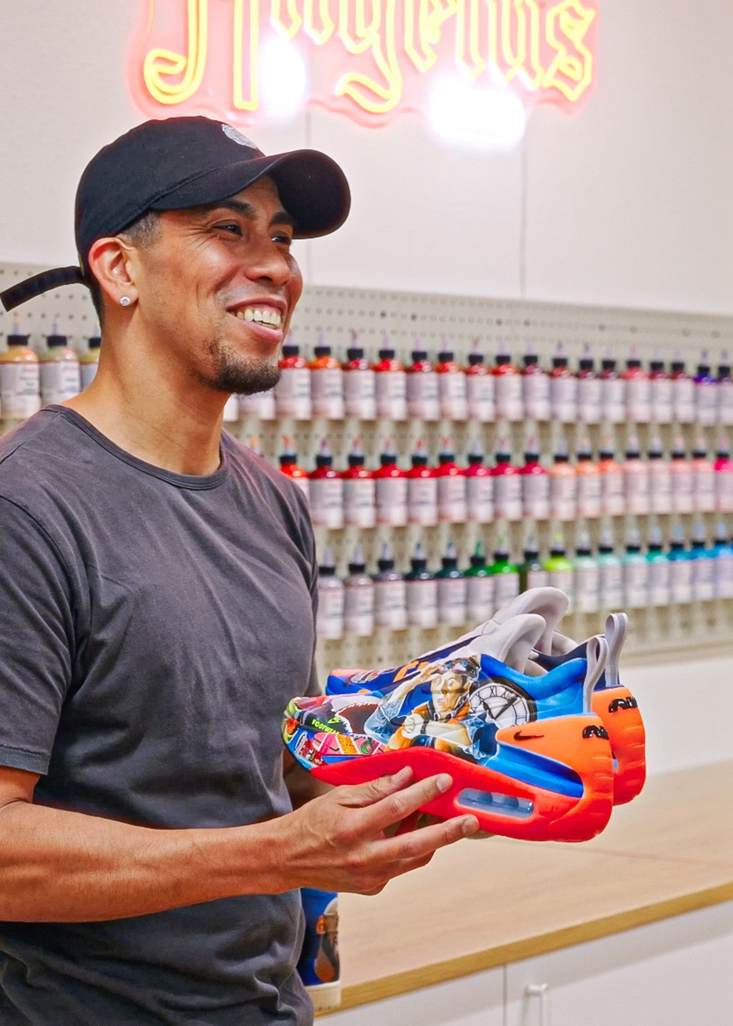 A smiling sneaker customizer in an Angelus workshop holding a pair of vibrant, hand-painted custom sneakers. In the background, a professional wall display features a full spectrum of Angelus Leather Paint bottles.