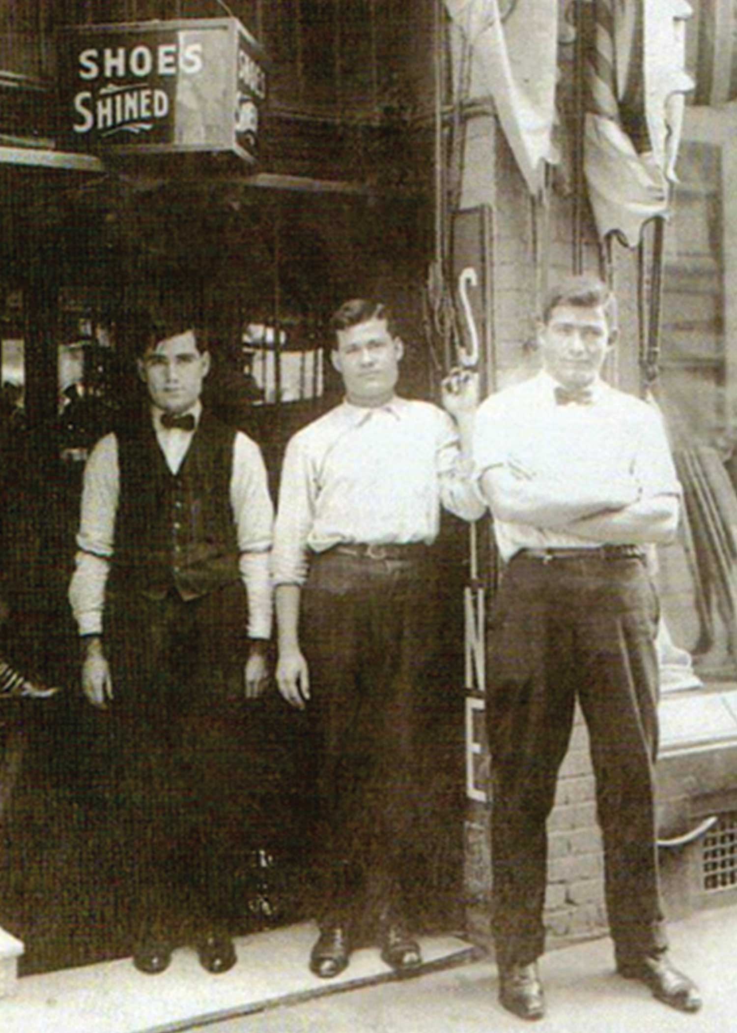 A vintage black-and-white photograph of Angelus Shoe Polish owners standing proudly in front of an early 20th-century 'Shoes Shined' storefront, representing the long-standing heritage of Angelus shoe care.