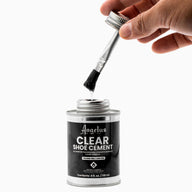A hand holds a brush applicator dipped in a 4 fl. oz. (118 ml) can of Angelus Direct Angelus Clear Shoe Cement, an all-purpose adhesive ideal for re-gluing shoe soles. The open silver can displays the product details on its label.