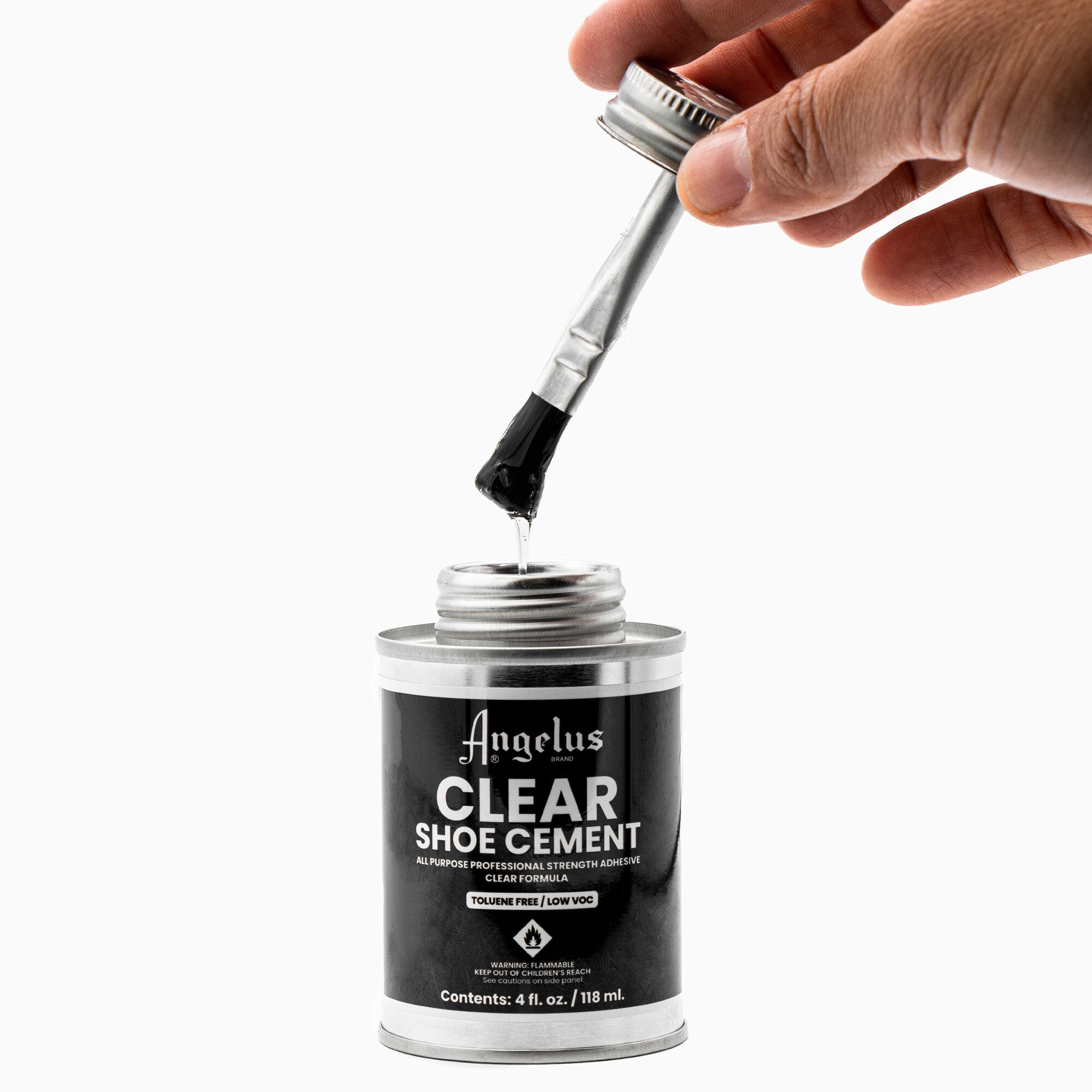 A hand holds a brush applicator dipped in a 4 fl. oz. (118 ml) can of Angelus Direct Angelus Clear Shoe Cement, an all-purpose adhesive ideal for re-gluing shoe soles. The open silver can displays the product details on its label.