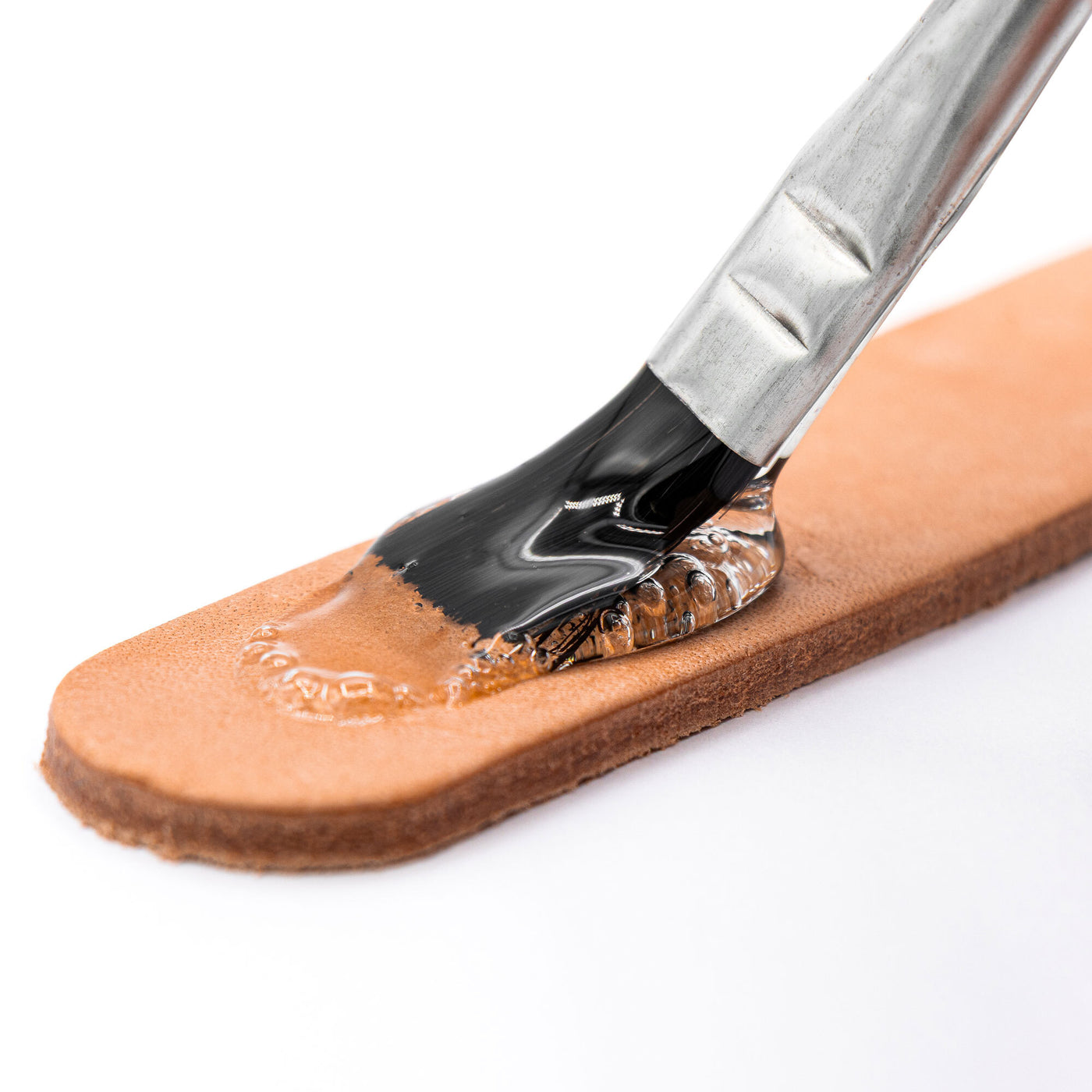 A close-up shows Angelus Direct’s Angelus Clear Shoe Cement being applied with a brush onto tan leather, ideal for repairs like re-gluing shoe soles—the brush is partially covered in the clear adhesive with small bubbles at the edge.