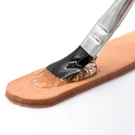 A close-up shows Angelus Direct’s Angelus Clear Shoe Cement being applied with a brush onto tan leather, ideal for repairs like re-gluing shoe soles—the brush is partially covered in the clear adhesive with small bubbles at the edge.