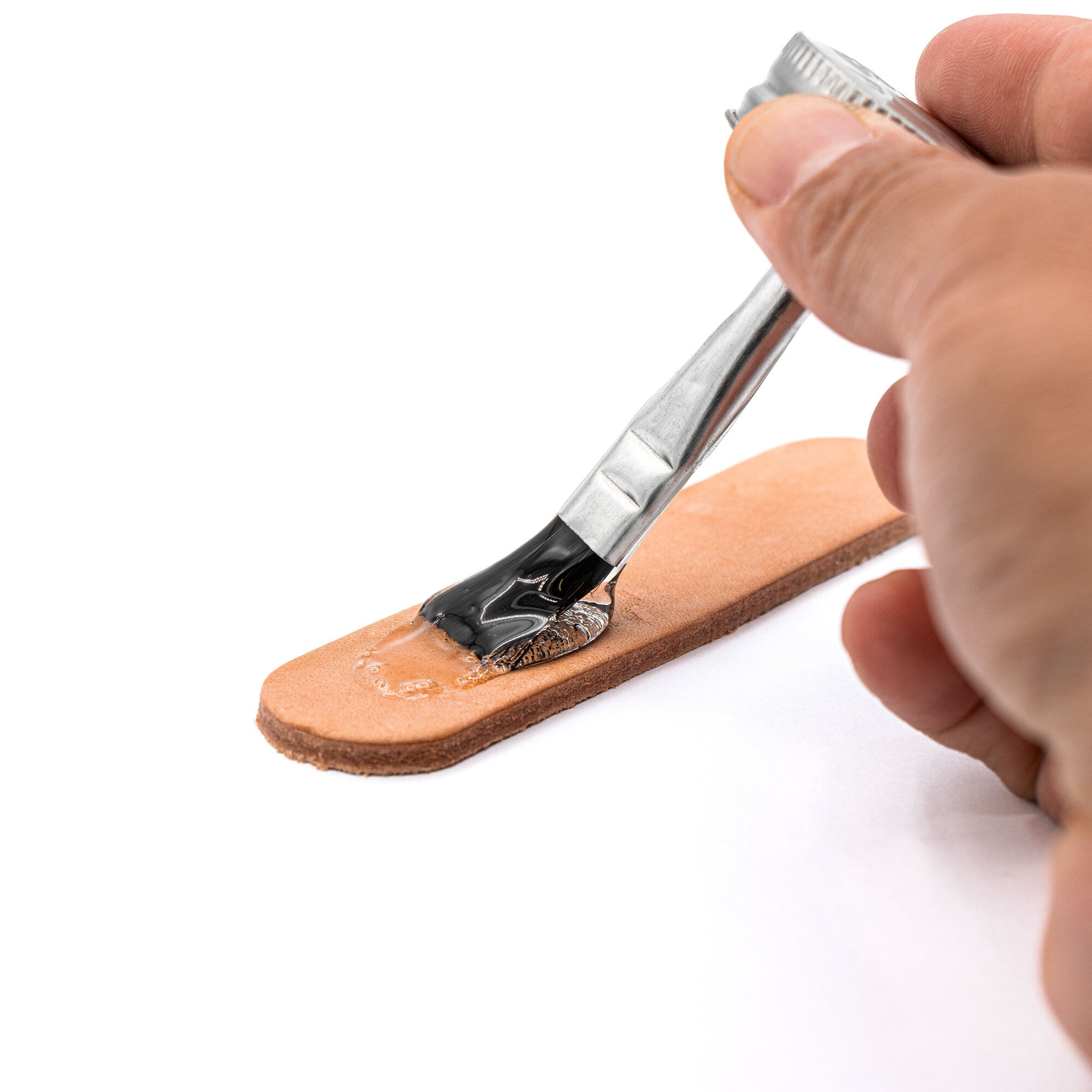 A hand uses a brush to apply Angelus Clear Shoe Cement by Angelus Direct onto a piece of brown leather on a white surface.