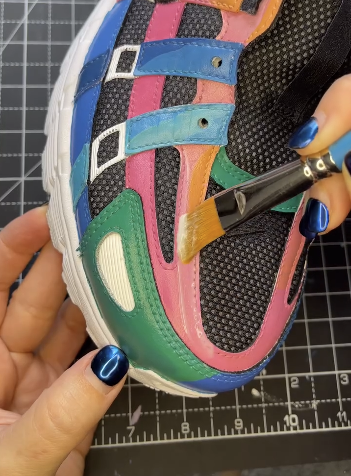 A flat brush applies a clear layer of Angelus Acrylic Finisher over a freshly painted pink leather section of a multi-colored custom sneaker to seal and protect the design.