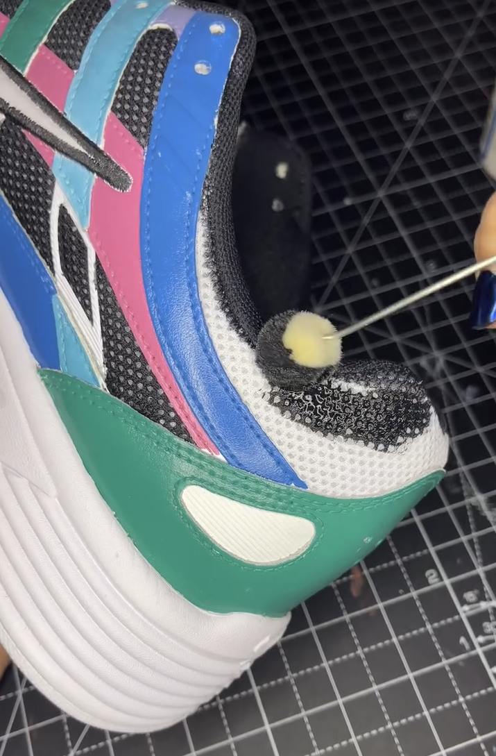 A hand using an Angelus wool dauber to apply black paint to the white mesh ankle collar of a colorful custom sneaker featuring vibrant teal, blue, and pink panels.