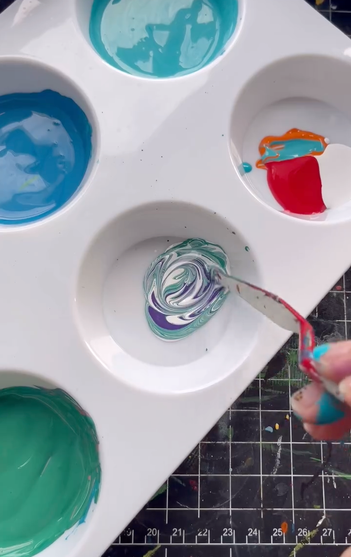 A close-up view of a white plastic paint palette where Angelus Leather Paints in teal, blue, and red are being mixed using a metal palette knife to create custom shades.