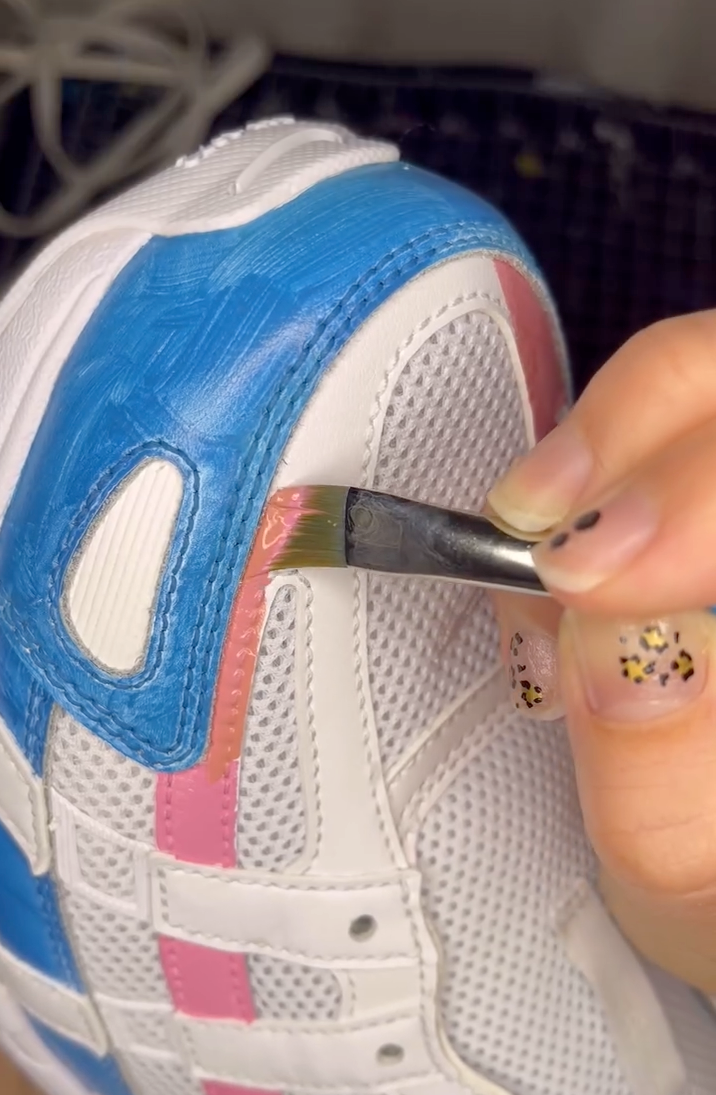 Close-up of a customizer applying pink Angelus Leather Paint to the leather overlays of a white mesh sneaker using a flat brush, with blue paint already applied to the heel.