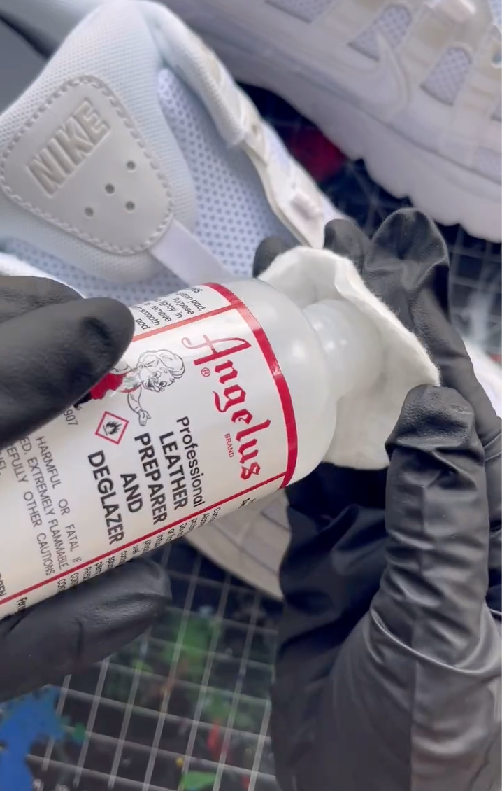A person wearing black nitrile gloves pours Angelus Professional Leather Preparer and Deglazer onto a cotton pad to prep a white Nike sneaker for painting on a gridded cutting mat.