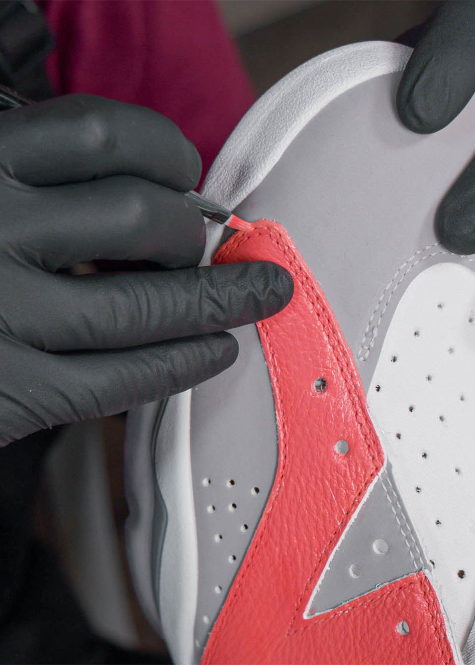 A detailed close-up of the 'Prep, Paint, Finish' process. A creator wearing black gloves uses a fine-detail brush to apply vibrant red Angelus Leather Paint to the textured leather panel of a grey and white sneaker.